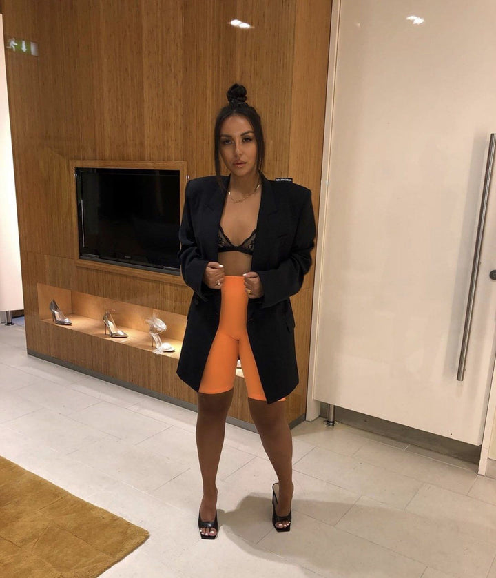 A woman wearing a black blazer, orange shorts, and black heels stands in a room with a wooden wall and a television.