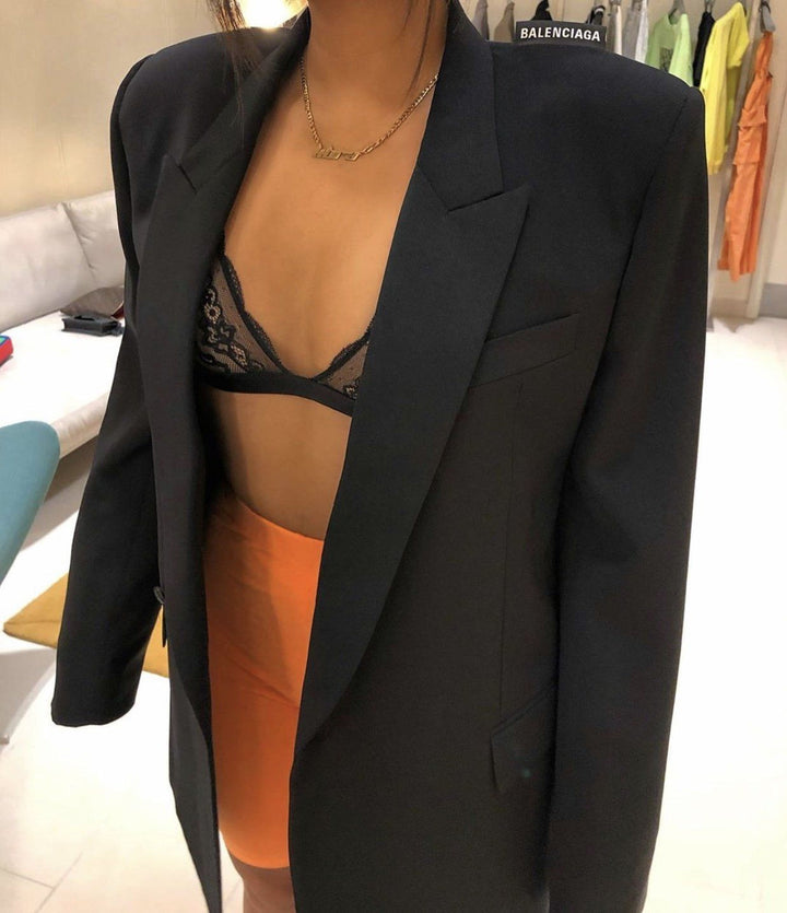 A woman wearing a black blazer, a black lace bra, and an orange skirt, standing in a clothing store.
