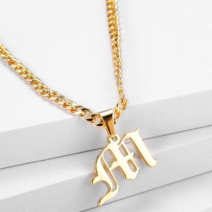 A gold-colored necklace with a pendant in the shape of the letter "M" on a white background.