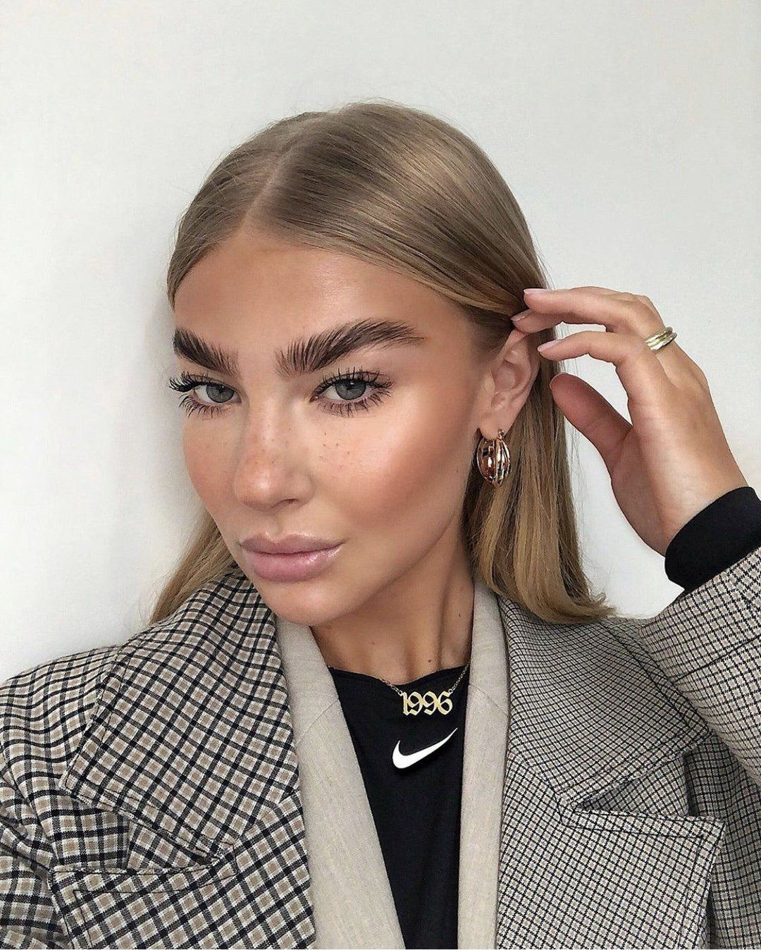 A woman with long blonde hair and a serious expression, wearing a black and white checkered jacket and a necklace with the year 1996.