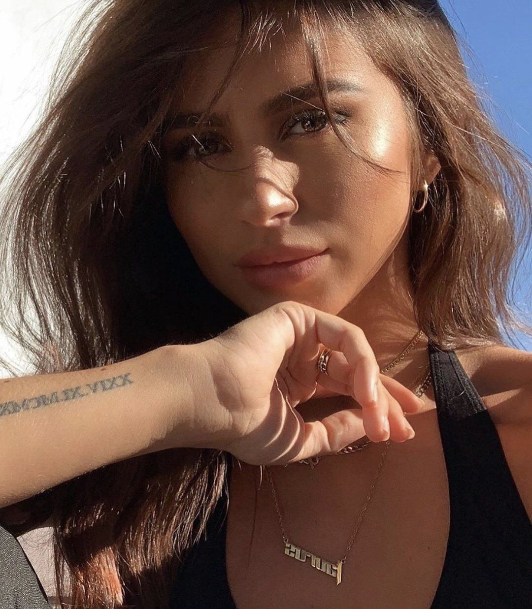 A close-up portrait of a young woman with long, dark hair and a thoughtful expression, wearing a black top and accessorized with a necklace and tattoos on her arm.