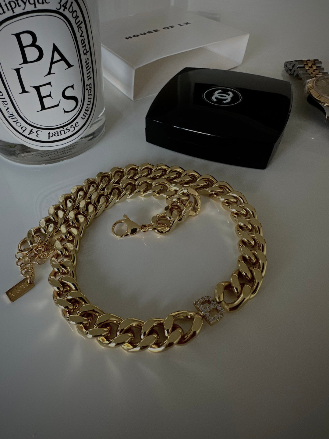 a gold chain necklace with a diamond-encrusted pendant, placed on a white surface alongside a black Chanel box and a clear glass bottle with a black label.