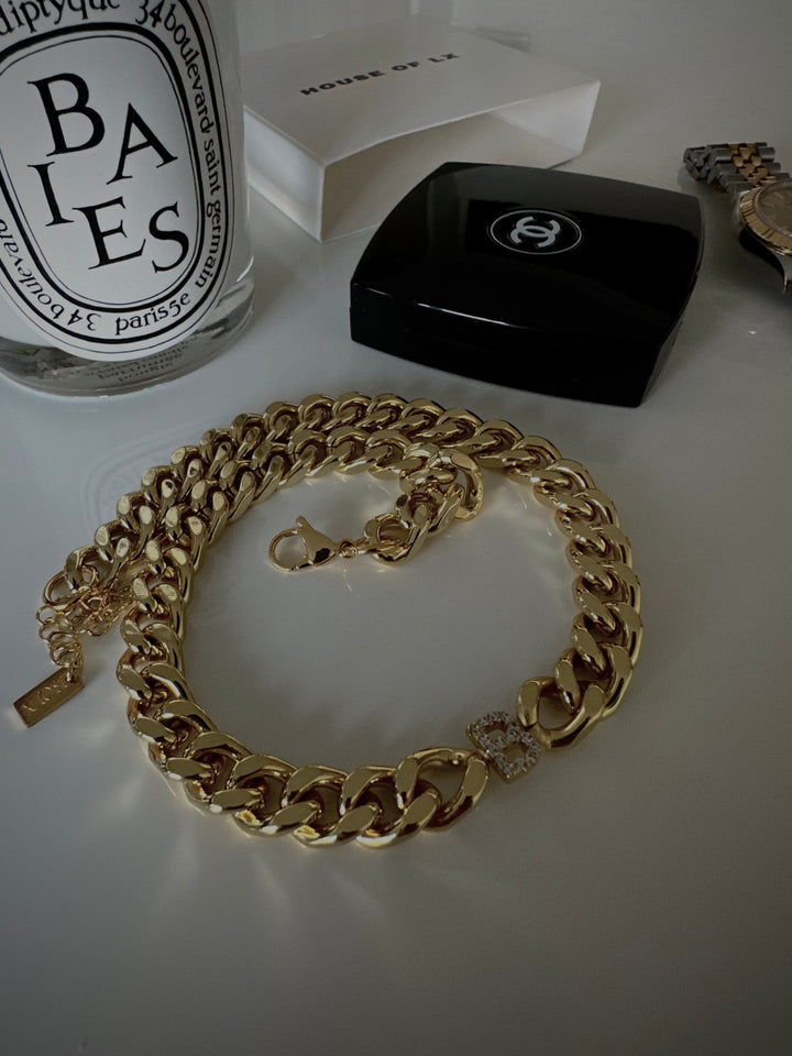 a gold chain necklace with a diamond-encrusted pendant, placed on a white surface alongside a black Chanel box and a clear glass bottle with a black label.